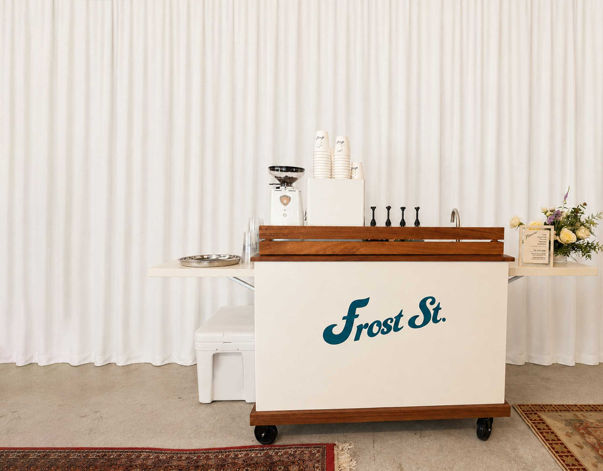 A white and wood Frost St. beverage cart with a teal logo, set against a white pleated backdrop with coffee equipment and cups on top.